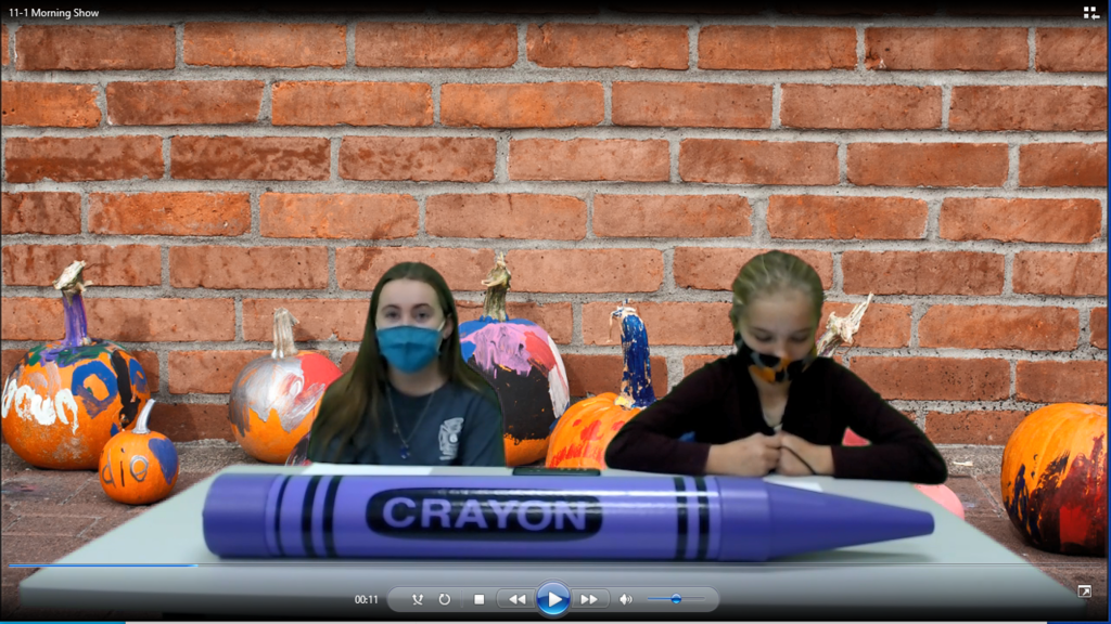 two girls sitting at a desk with a giant purple crayon on it.