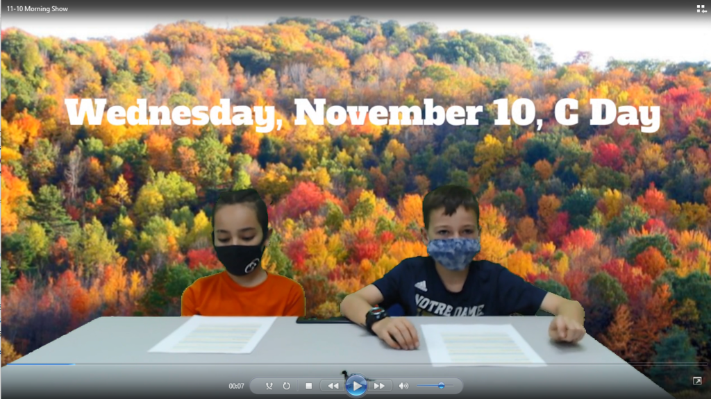 two students at news desk with colorful fall  trees in background